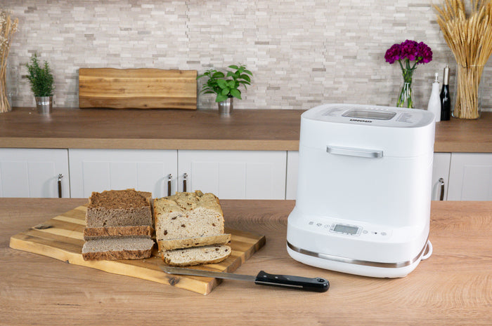 Der Brotbackautomat der Unold AG Backmeister Bernd steht auf einem Kรผchentisch neben einem hรถlzernen Schneidebrett mit verschiedenen Brotscheiben und einem Brotmesser; im Hintergrund sind Pflanzen, Blumen und ein weiteres Schneidebrett zu sehen.