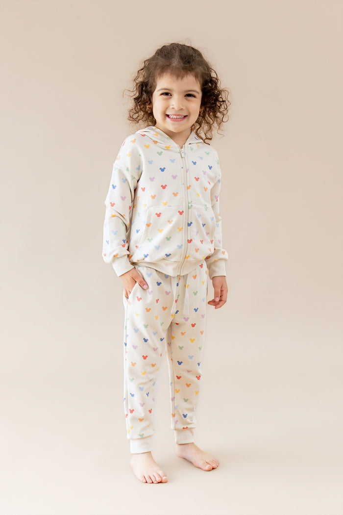 Child wearing a mickey-patterned zip up hoodie and jogger pant on a beige background