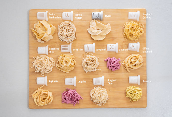 Die Unold Nudelmaschine Nora verfรผgt รผber 15 Nudelsorten, jede mit deutscher Beschriftung und den entsprechenden Aufsรคtzen auf einem Holzbrett angeordnet, und zeigt Formen wie Spaghetti, Fettuccine, Ravioli und Makkaroni.