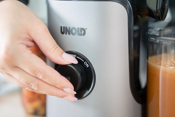 Eine Hand mit manikรผrten Nรคgeln dreht den Regler am silbernen Unold Slow Juicer Sam von AUS auf EIN, daneben steht ein Glas orange-braunen Safts.