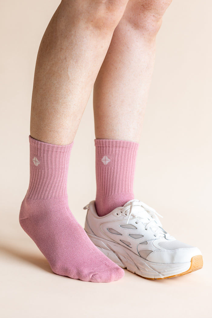 A close-up of a person's feet wearing pink crew socks and a white athletic sneaker against a solid light beige background.