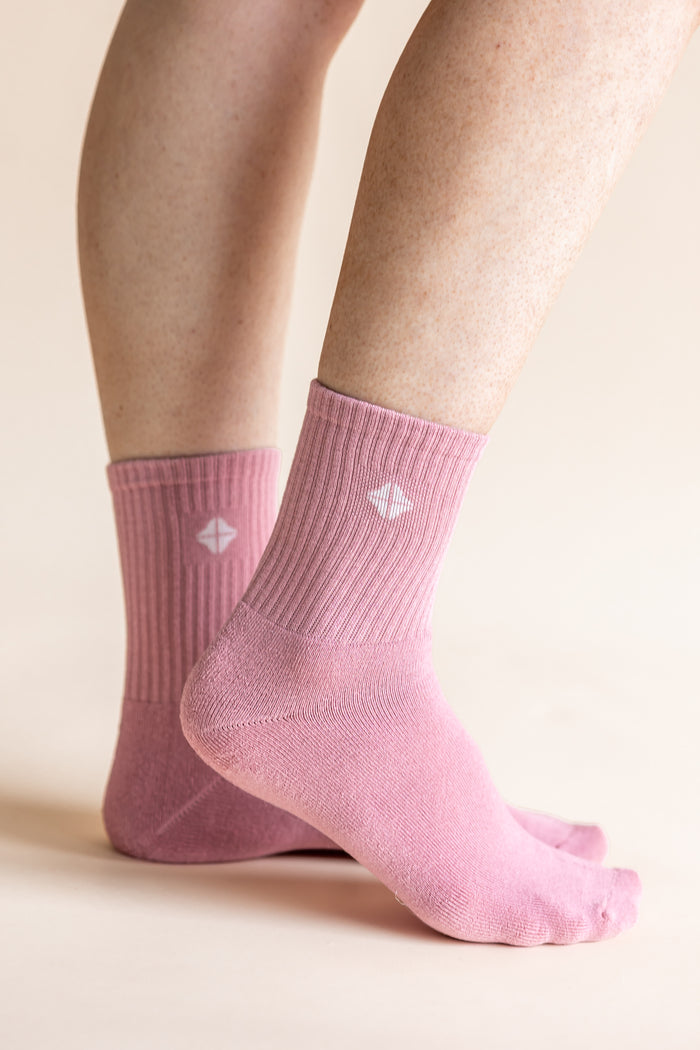 A close-up of a person's feet wearing light pink ribbed crew socks with a small white geometric logo on the side, set against a solid pale beige background.