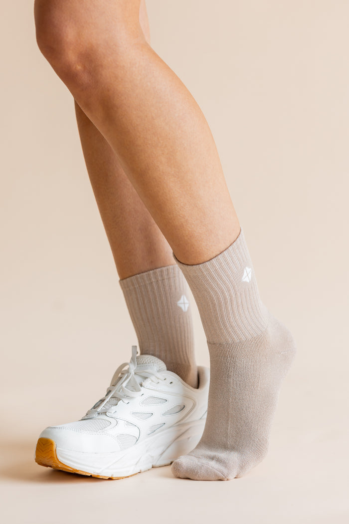 A close-up of a person's legs wearing light beige ribbed crew socks and a white athletic sneaker against a solid beige background.