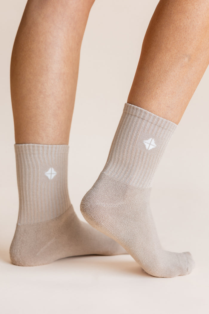 A close-up of a person's feet and lower legs wearing light beige crew socks with a small white geometric logo on the side, set against a solid warm beige background.
