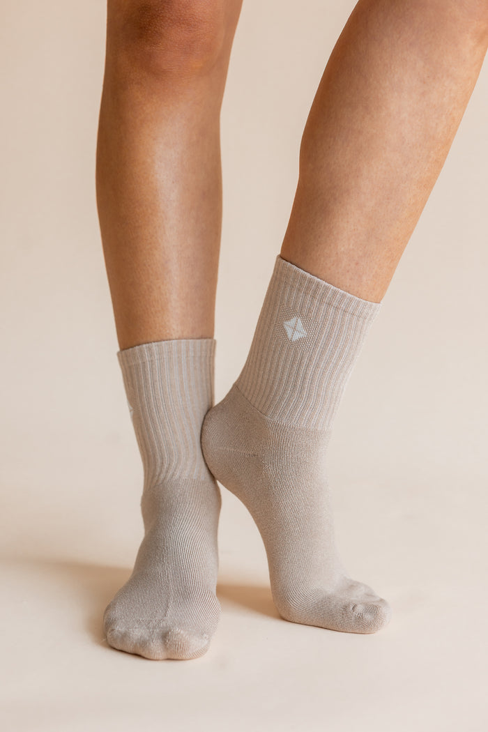 A close-up of a person's feet and lower legs wearing light gray ribbed crew socks against a warm beige studio background.