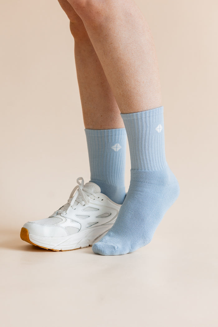 A person's legs and feet wearing light blue crew socks and a white athletic sneaker, shown against a solid pale beige background.