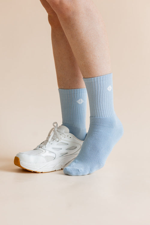 A person's legs and feet wearing light blue crew socks and a white athletic sneaker, shown against a solid pale beige background.