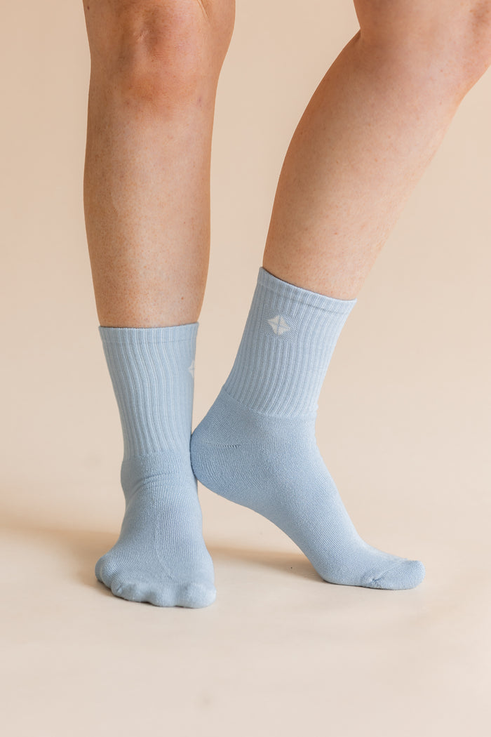A pair of light blue ribbed crew socks worn on a person's feet, shown against a solid beige background.