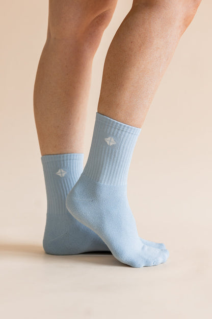 A pair of light blue crew socks worn on a person's feet, shown against a plain beige studio background. A small white geometric logo is visible on each sock.