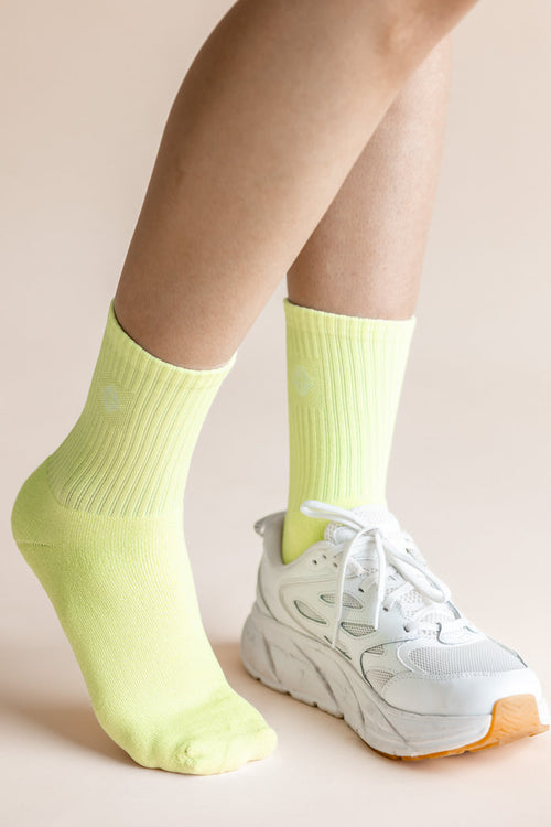 A close-up of a person's legs wearing neon yellow ribbed crew socks and a white chunky sneaker against a solid pale beige background.