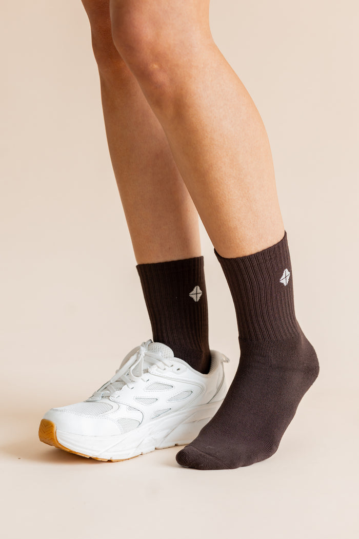 A close-up of a person's legs wearing dark brown ribbed crew socks and a white athletic sneaker against a plain beige studio background.