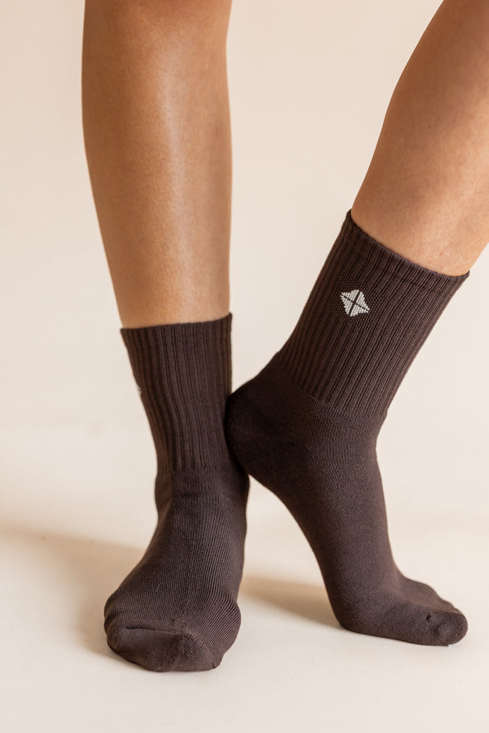 A close-up of a person's feet wearing dark brown ribbed crew socks against a warm beige background. A small white geometric logo is visible on the side of the right sock.