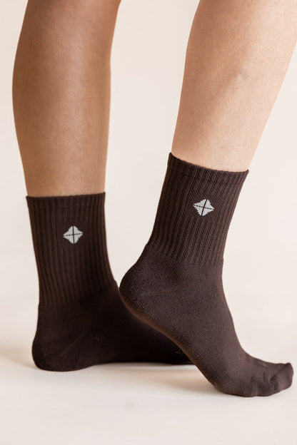 A close-up of a person's feet wearing dark brown crew socks with a small white geometric logo on the side, set against a plain light beige background.