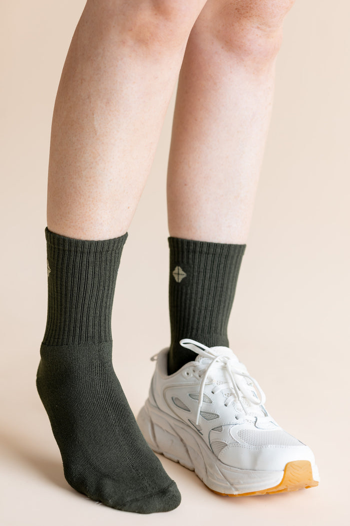A close-up of a person's lower legs wearing a pair of dark charcoal gray ribbed crew socks and a white chunky sneaker against a solid beige background.