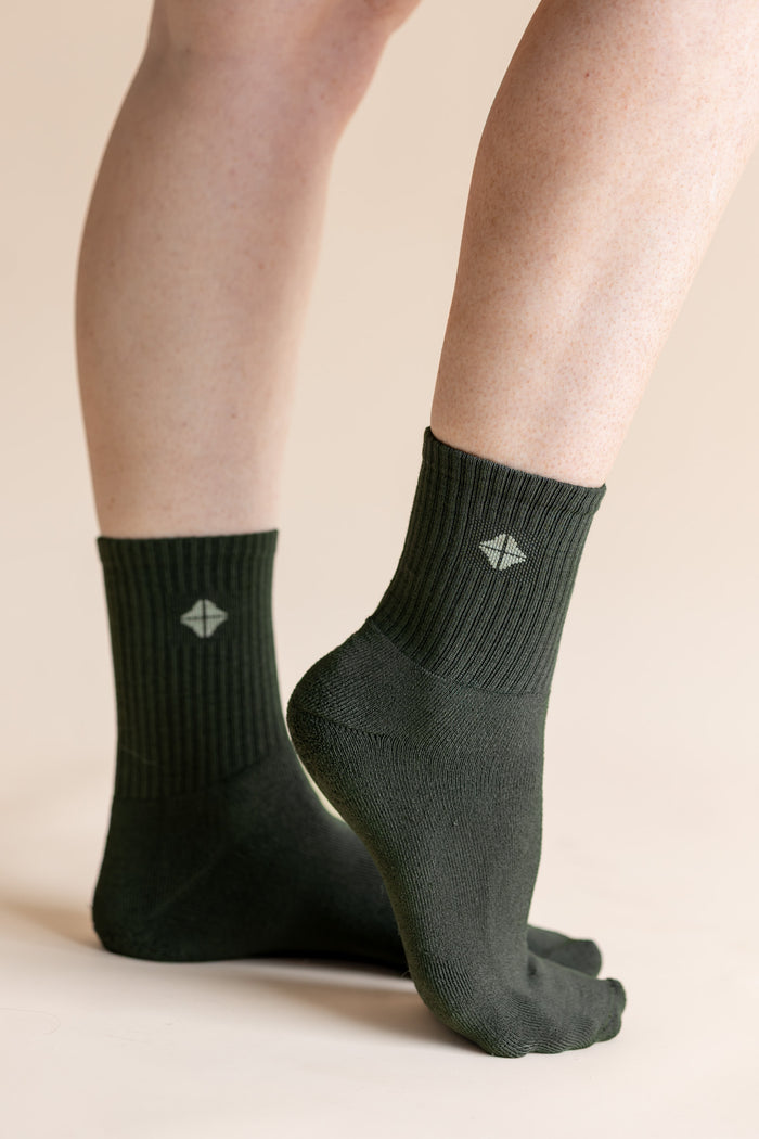 A pair of dark forest green crew socks worn on a person's feet, shown against a solid light beige background. The socks feature a ribbed cuff and a small white geometric logo on the front.