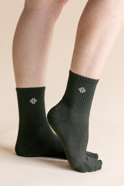 A pair of dark forest green crew socks worn on a person's feet, shown against a solid light beige background. The socks feature a ribbed cuff and a small white geometric logo on the front.