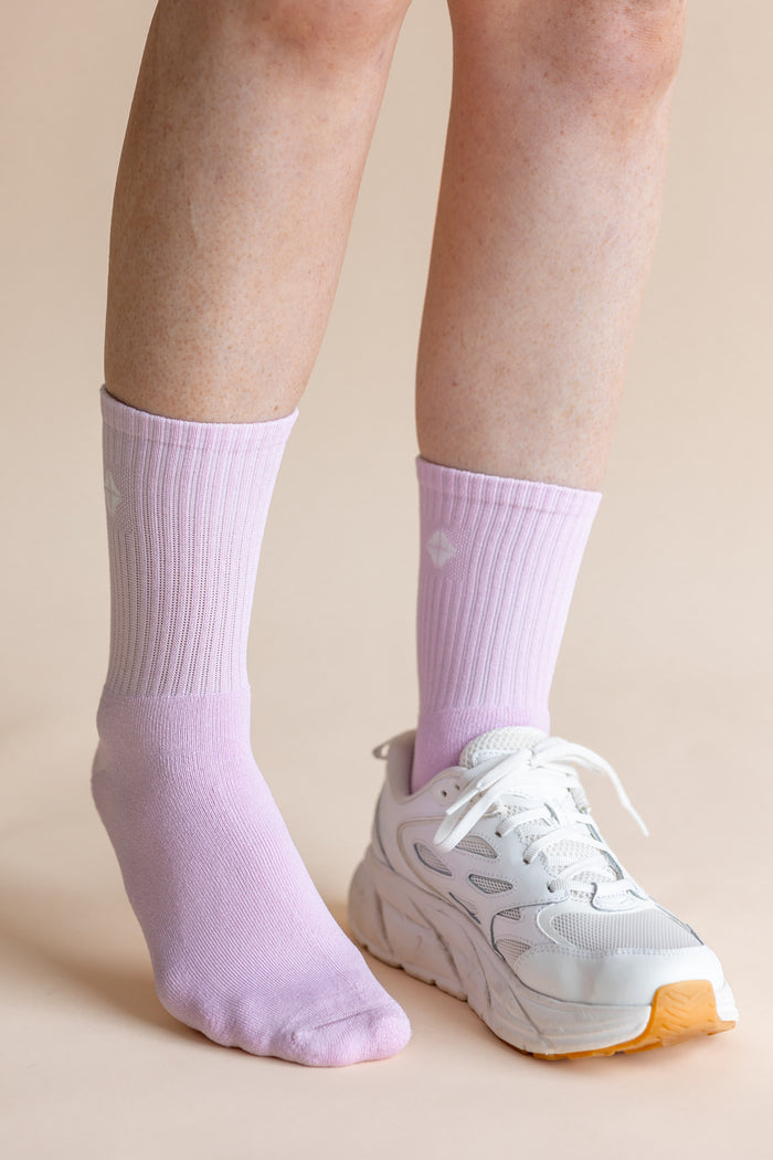 A close-up of a person's lower legs wearing light purple ribbed crew socks and a white chunky sneaker against a solid beige background.