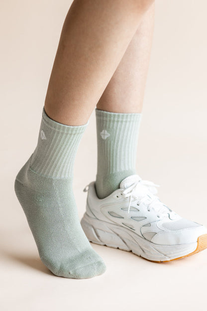 A close-up of a person's feet wearing pale sage green ribbed crew socks and white athletic sneakers against a soft beige background.