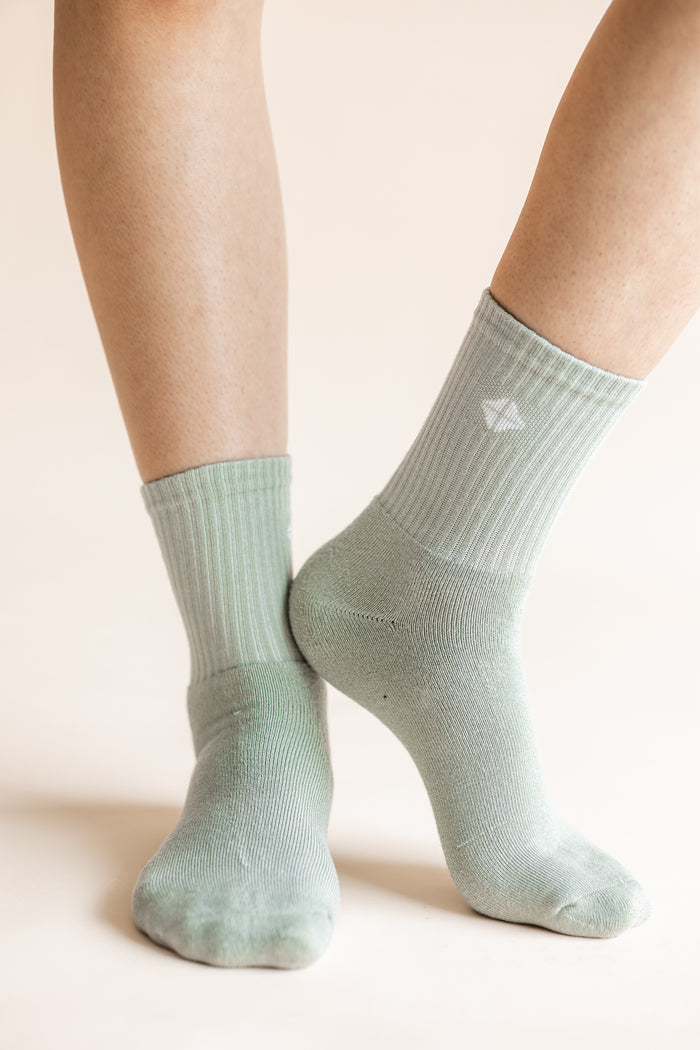 A close-up of a person's feet wearing ribbed crew socks in a pale sage green color, with a small white geometric logo on the side of the right sock.