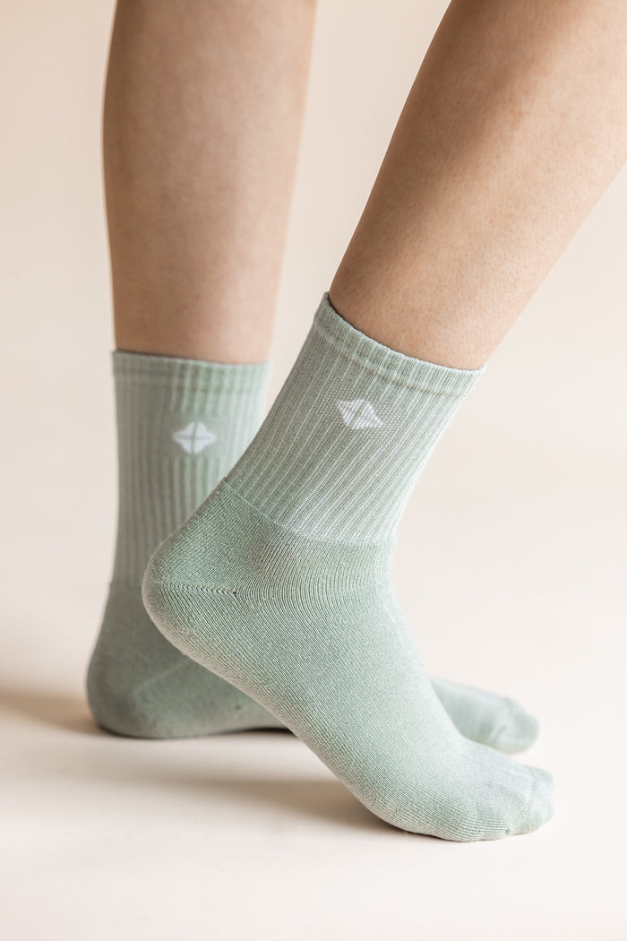 A close-up of a person's feet wearing light sage green ribbed crew socks with a small white geometric logo on the side, set against a warm beige background.