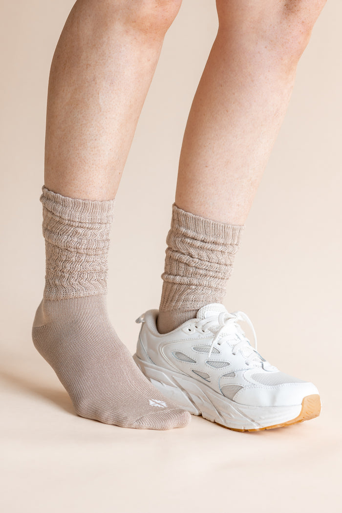A close-up of a person's legs wearing beige ribbed crew socks and a white athletic sneaker against a solid light beige background.