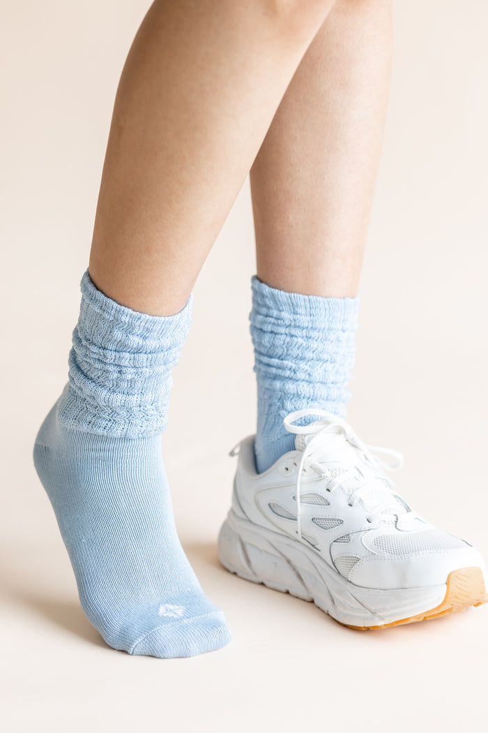 A close-up of a person's legs wearing light blue ribbed crew socks and a white athletic sneaker against a plain beige background.