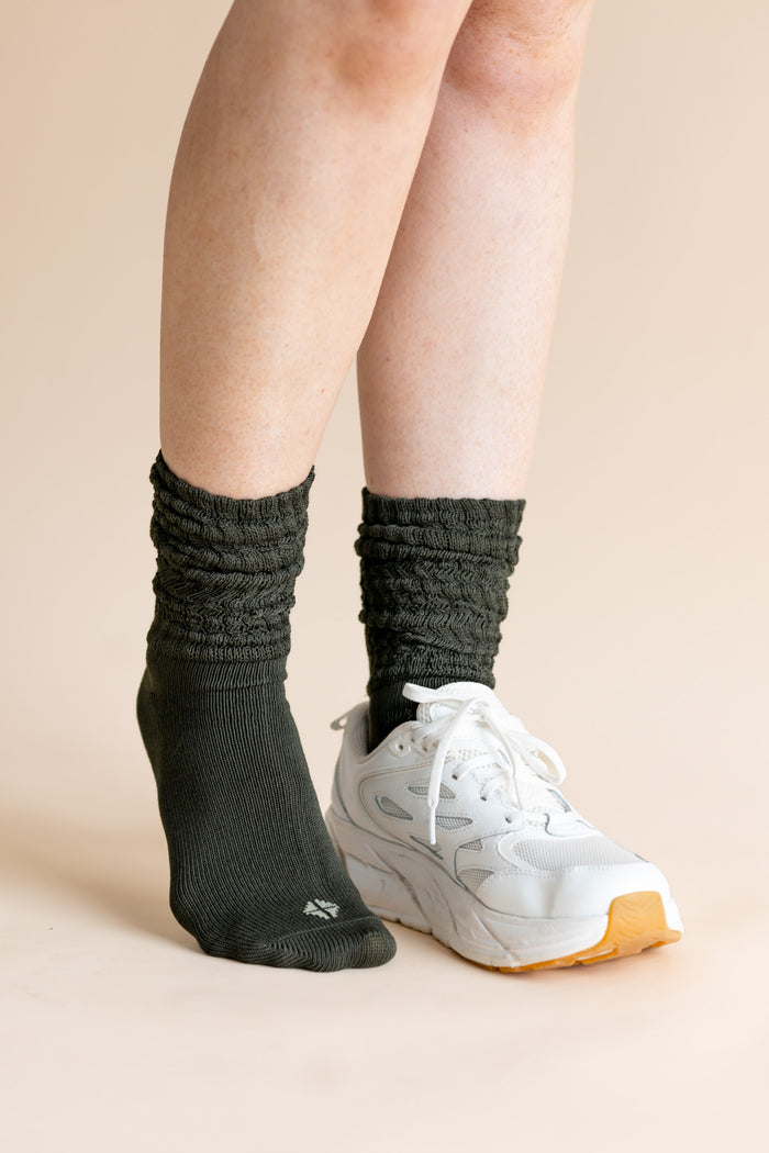A person's legs wearing dark charcoal gray ribbed crew socks and a white athletic sneaker, set against a solid light beige background.