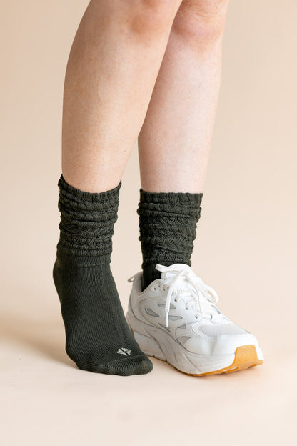 A person's legs wearing dark charcoal gray ribbed crew socks and a white athletic sneaker, set against a solid light beige background.
