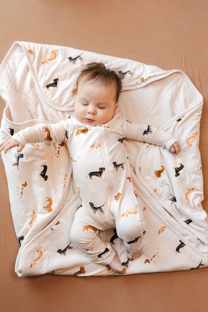 Baby sleeping over a beige Blanket with Dachshund print wearing a similar print romper on a brown background