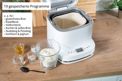 Der Brotbackautomat der Unold AG Backmeister Bernd steht offen auf einem Holztisch, umgeben von Mehl, Wasser, Hefe und einem Löffel. Ein Schild weist auf seine 19 voreingestellten Programme für verschiedene Brote und Teige hin.