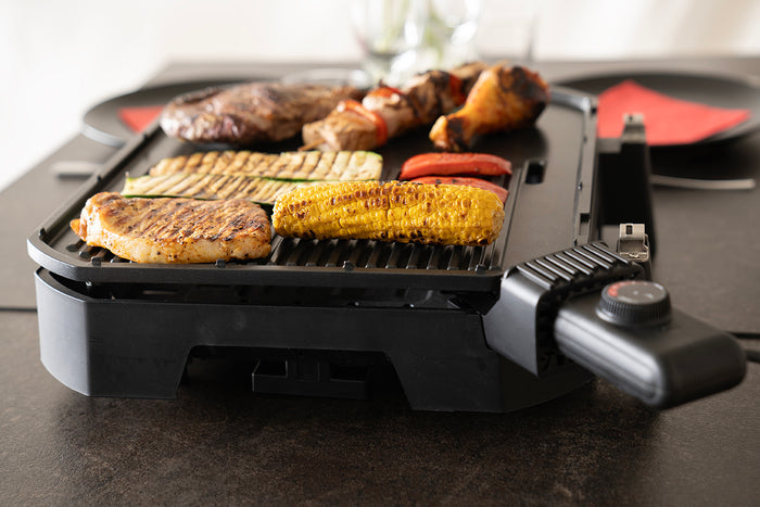 Der Unold BBQ-Stand-/Tischgrill Jimmy, ein schwarzer Elektrogrill mit Temperaturregler am Griff, steht auf einem Tisch und grillt Maiskolben, Hähnchenbrust, Steak, Spieße, Zucchini und Tomatenscheiben.