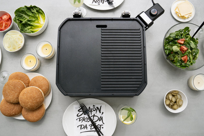Eine Draufsicht auf den Elektrogrill Unold BBQ-Stand-/Tischgrill Jimmy auf einem Leuchttisch, umgeben von Burgerbrötchen, Salat, Zwiebeln, Tomatenscheiben, Essiggurken, Käse, Salat, Kerzen, Getränken und Tellern.