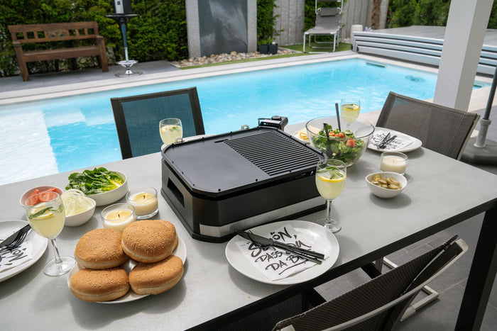 Ein Terrassentisch am Pool ist mit Burgern, Gemüse, Salaten, Weingläsern, Tellern und dem Elektrogrill Unold BBQ-Stand-/Tischgrill Jimmy gedeckt und bereit für eine Mahlzeit im Freien.