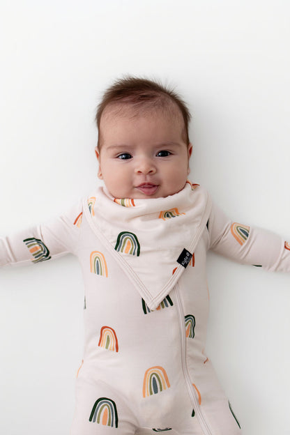 Baby wearing Bib in Retro Rainbow print over a zippered romper with similar print on a white background