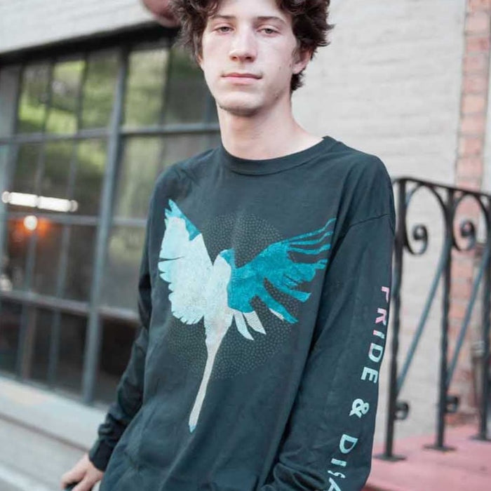 A young person with short brown hair stands outdoors by a window and staircase, wearing a Sleep On It long-sleeve T-shirt featuring a large bird graphic and "PRIDE & DISGRACE" printed on the sleeve.