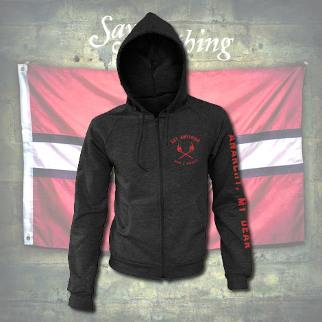 A black Say Anything Cotton Heritage zip-up hoodie with red crossed axes and chest text, plus red writing on the right sleeve, is shown in front of a red-white-red striped flag on a textured wall.