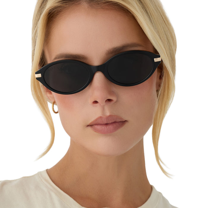 on model - blonde female wearing diff eyewear claudine oval sunglasses with a black acetate frame, gold metal temples and grey polarized lenses front view