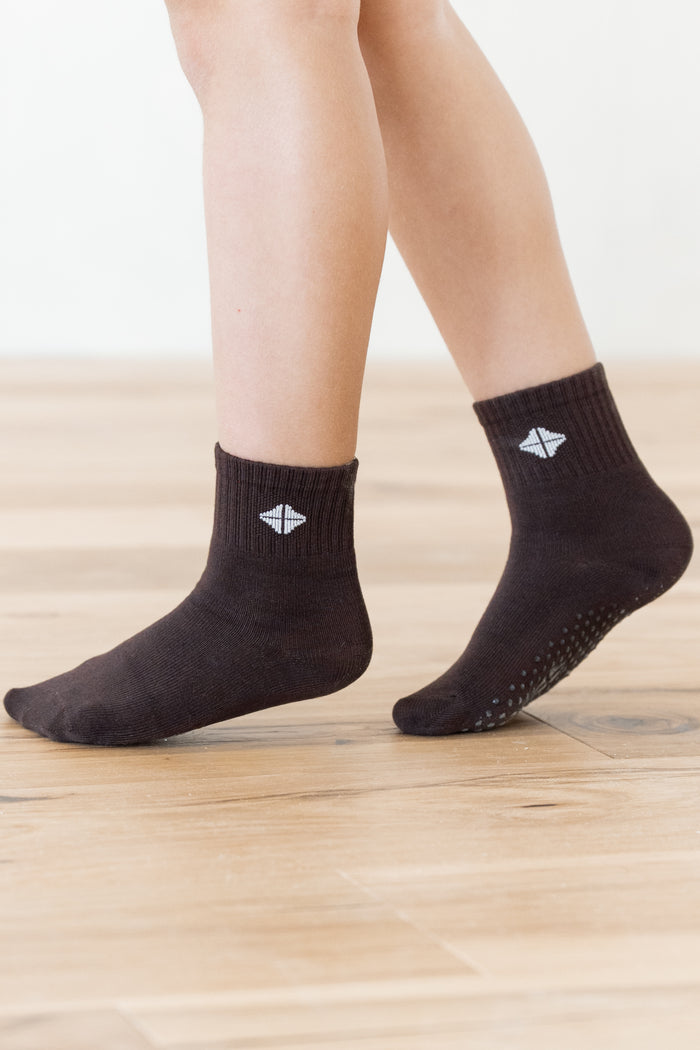 A pair of dark brown ankle socks worn on a person's feet, standing on a light wood floor against a white wall. One sock has a small white geometric logo, and the sole of the other sock shows grip dots.