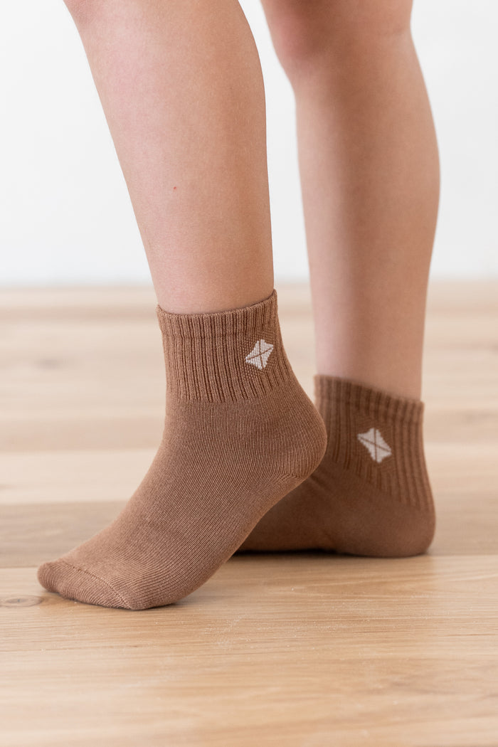 A close-up of a person's feet wearing brown ankle socks with a small white geometric logo on the side, standing on a light wood floor against a white wall.