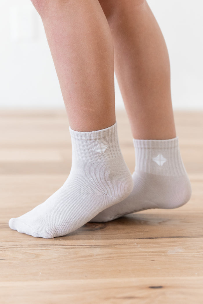 White ankle socks worn on a child's feet, shown from the calves down on a light wood floor.