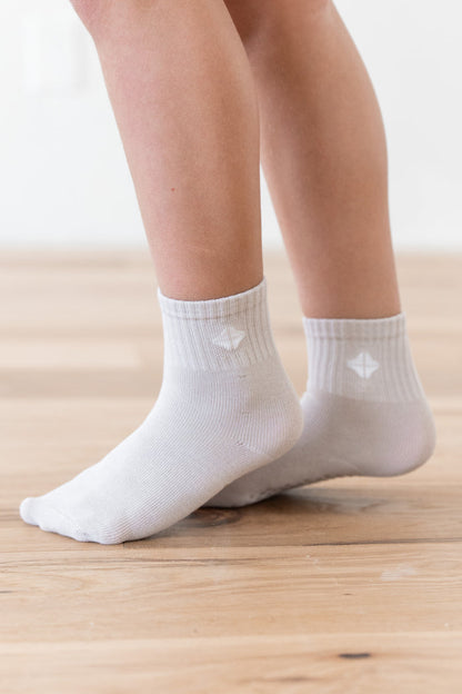 White ankle socks worn on a child's feet, shown from the calves down on a light wood floor.