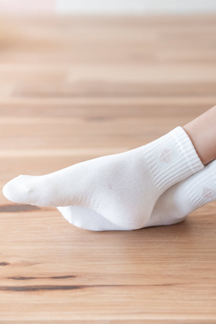 A close-up of a person's foot wearing a white ankle sock, standing on a light brown wooden floor.