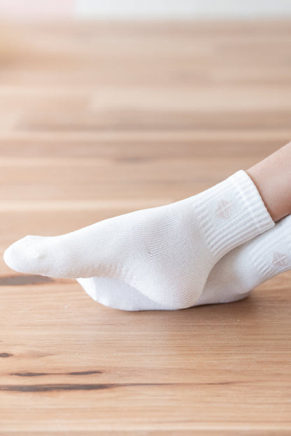 A close-up of a person's foot wearing a white ankle sock, standing on a light brown wooden floor.