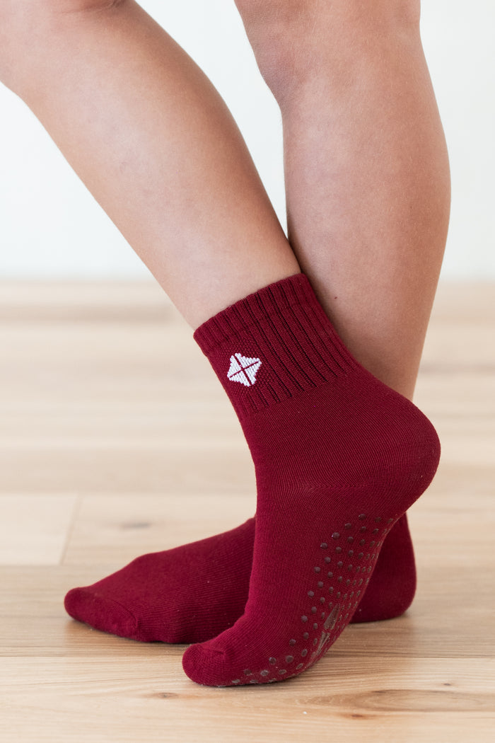 A close-up of a person's feet wearing dark red ankle socks with grip dots on the sole and a small white geometric logo on the side, standing on a light wood floor.