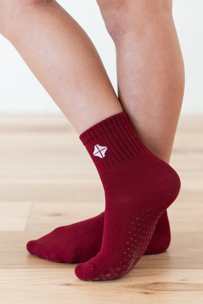 A close-up of a person's feet wearing dark red ankle socks with grip dots on the sole and a small white geometric logo on the side, standing on a light wood floor.