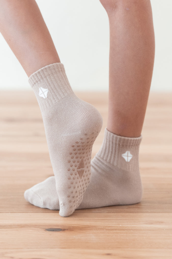 A close-up of a person's feet wearing light beige ankle socks with textured grip dots on the sole and a small white geometric logo near the cuff, standing on a light wood floor.