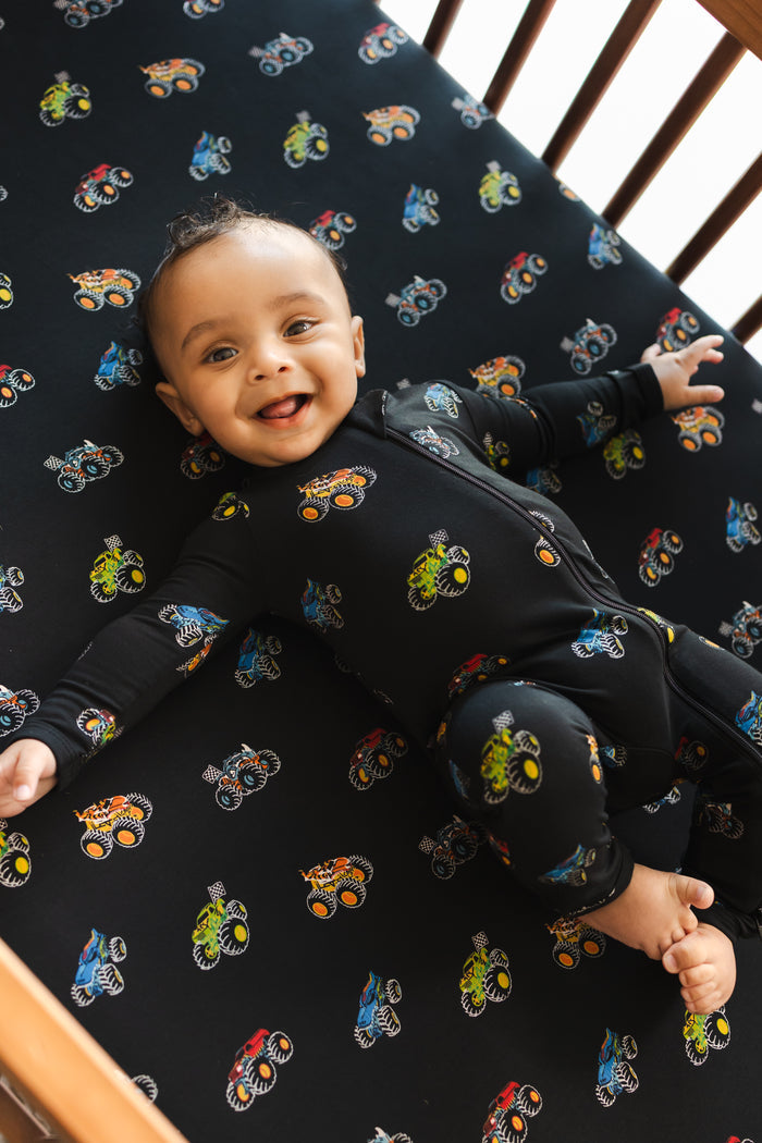 Baby lying on black crib sheet in Monster Truck print wearing similar print Zippered Romper