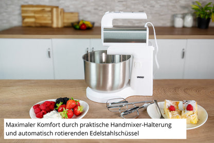 Der Unold Handmixer Hannah mit Rührschüssel, ein weißer Standmixer mit Edelstahlschüssel, steht auf der Küchentheke neben Tellern mit Beeren und Kuchen. Der deutsche Text hebt seinen Komfort, den praktischen Handmixerhalter und die automatisch rotierende Schüssel hervor.