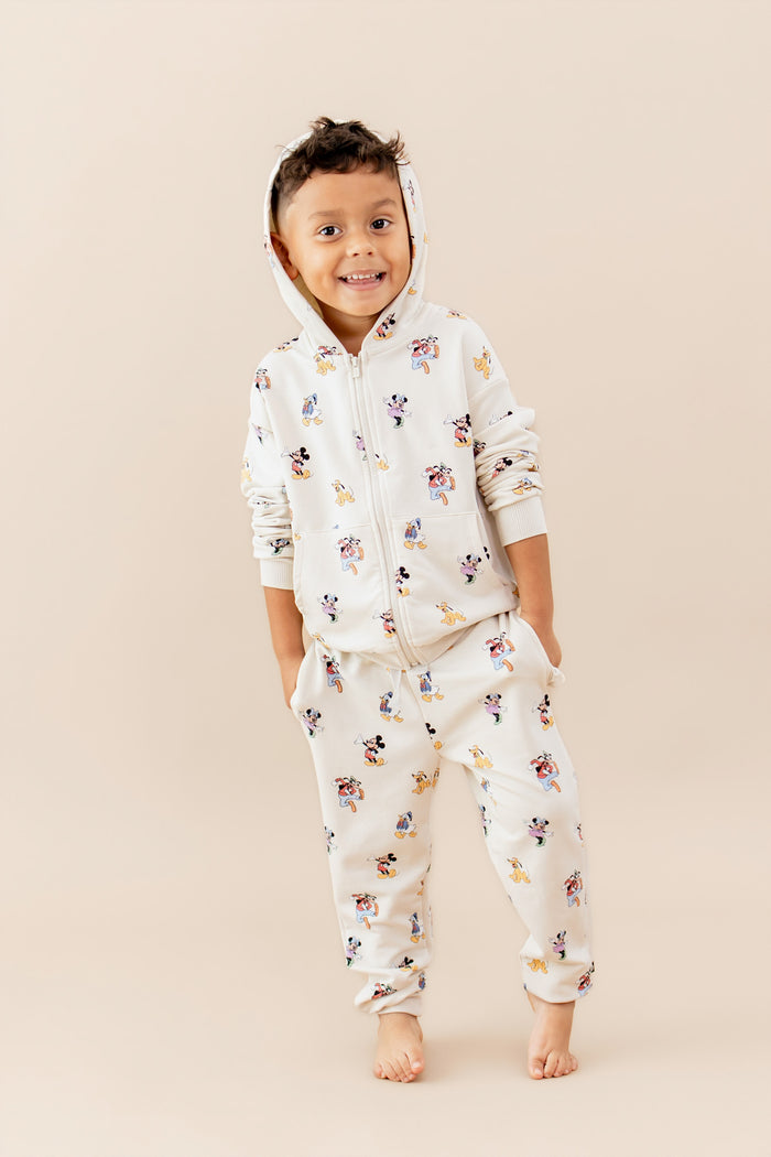 Child modeling a matching sweatsuit with a mickey and friends pattern featuring a zip up hoodie and jogger style pants