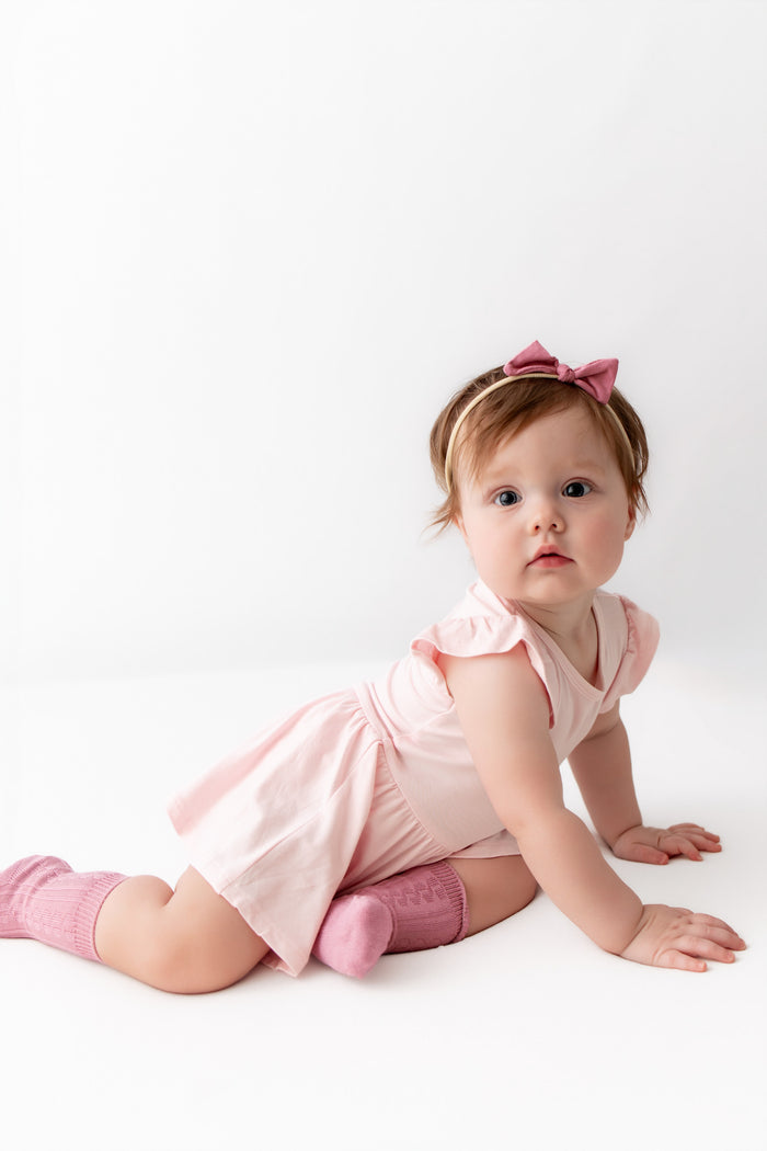 A baby girl crawling on a white studio floor, wearing a light pink ruffle-sleeve dress, pink socks, a pink bow headband, and looking directly at the camera.